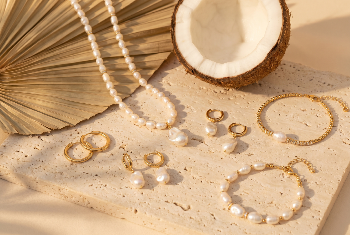 Flat lay of gold and pearl jewelry on travertine stone with coconut half and palm frond shadow
